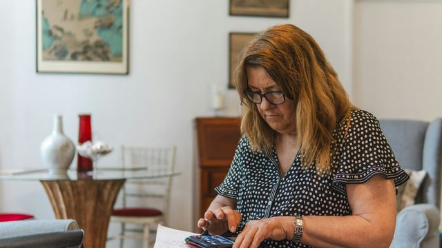 Mulher de óculos sentada na poltrona de casa, fazendo contas na calculadora com papéis no colo