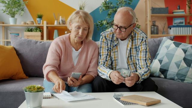 Casal de idosos sentado em um sofá analisando contas e papéis, ela com o celular na mão e ele segurando notas de dinheiro.