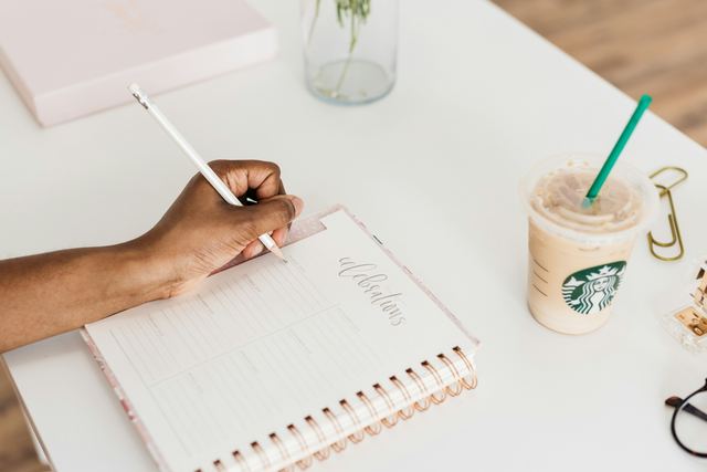 Mão escrevendo numa agenda aberta sobre a mesa, ao lado de um café gelado