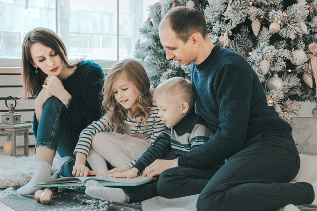 Família lendo um livro no chão, junto à árvore de Natal, com as duas crianças entre os pais