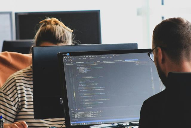 Duas pessoas programando diante de monitores com código num escritório claro