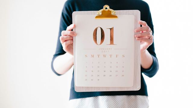 Pessoa de blusa escura segurando uma prancheta com o calendário de janeiro à frente do corpo, com as duas mãos à vista