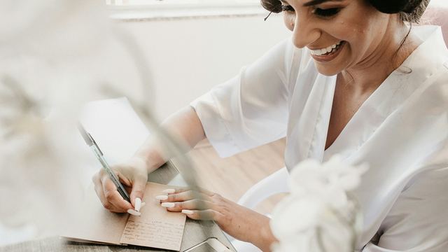 Mulher de blusa branca sorrindo enquanto escreve numa agenda de papel, com o celular ao lado na mesa