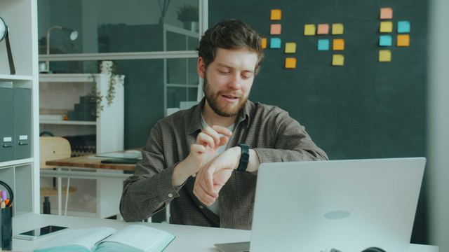 Homem de camisa marrom sentado em uma mesa de escritório com notebook, olhando o smartwatch no pulso.