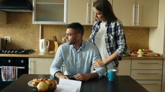Homem sentado à mesa da cozinha olhando contas enquanto a esposa, de pé atrás dele, o abraça pelos ombros.