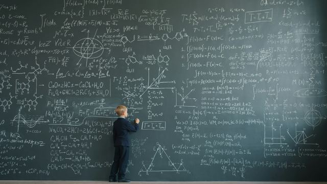 Menino de terno escrevendo fórmulas matemáticas em uma lousa grande de sala de aula