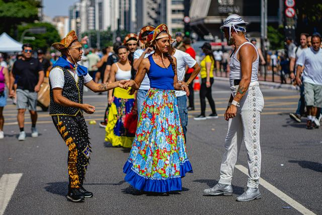 Pessoas com fantasias coloridas dançando e se apresentando em uma rua fechada, com prédios ao fundo.