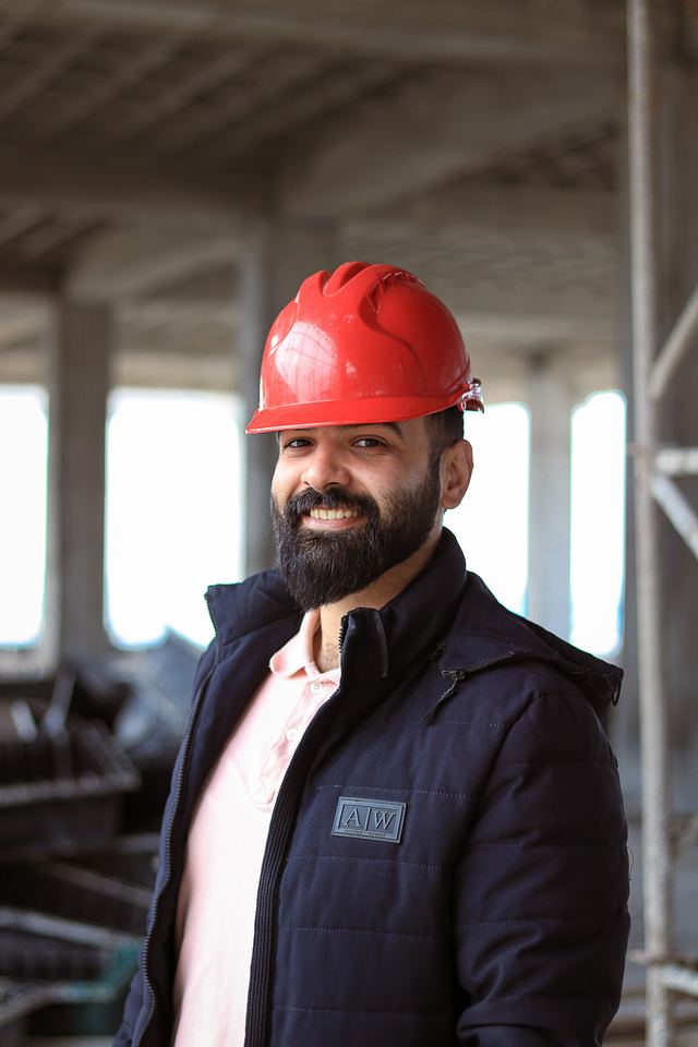 Homem de barba sorrindo, com capacete vermelho e jaqueta azul-marinho, dentro de uma obra em construção