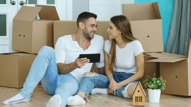 Casal sentado no chão da casa nova, entre caixas de mudança, olhando um tablet juntos