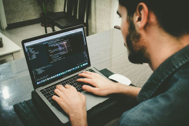 Homem de barba, visto de perfil, digita código em um notebook sobre uma mesa.