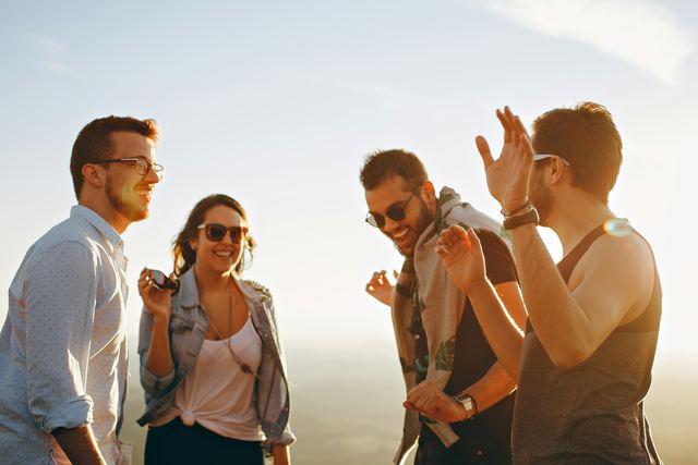 Quatro amigos, três homens e uma mulher, rindo juntos ao ar livre num dia de sol.