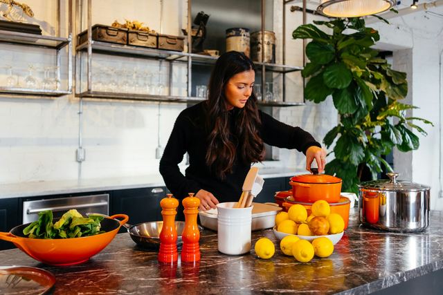 Mulher numa cozinha segurando a tampa de uma panela diante de uma bancada com frutas