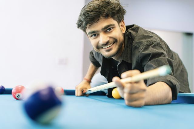 Jovem sorrindo enquanto mira a bola com o taco numa mesa de sinuca