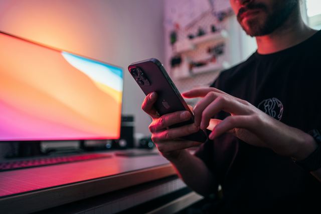 Homem de camiseta preta segurando um celular diante da mesa de trabalho