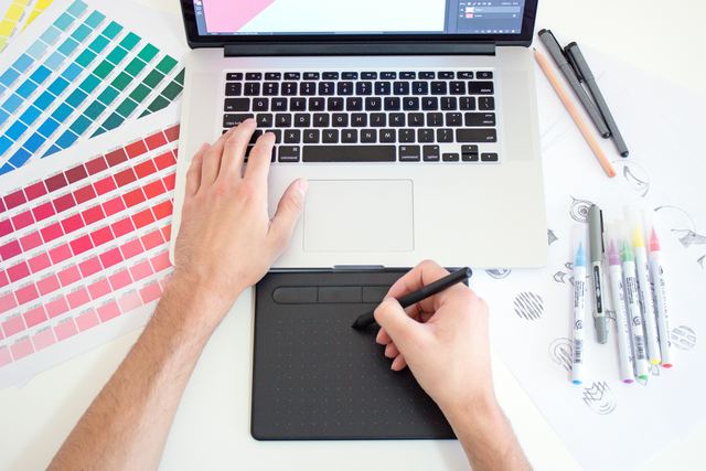 Designer de gráficos trabalhando em um laptop com mesa digitalizadora, amostras de cor e canetas na mesa