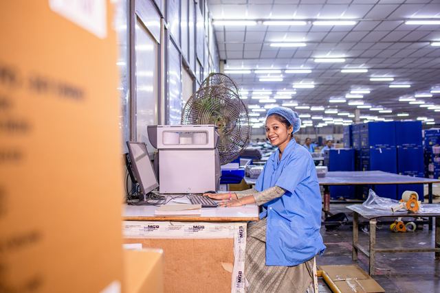 Funcionária de uniforme azul e touca sorri sentada em frente a um computador em um galpão de embalagem.