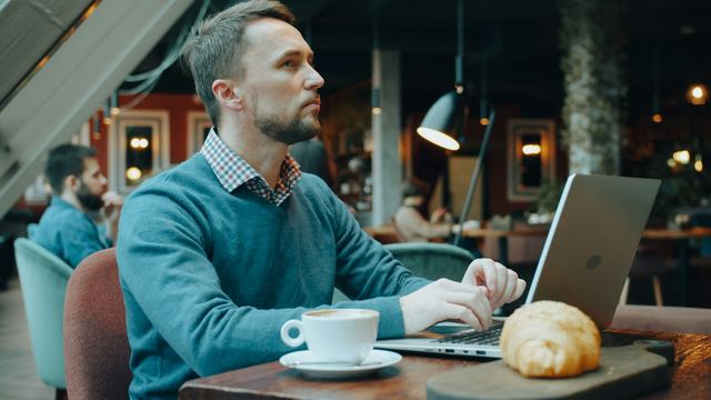 Homem de suéter azul digitando no notebook numa mesa de café