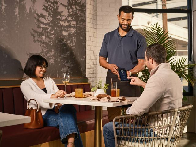 Garçom em pé mostrando uma maquininha a um casal sentado à mesa de um restaurante, enquanto o homem toca a tela.