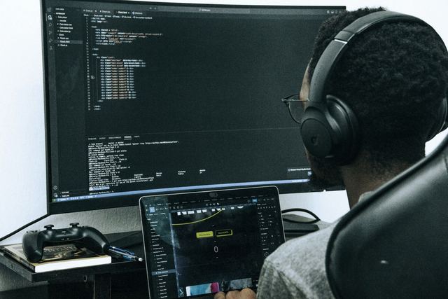Homem de óculos e fones de ouvido, visto de lado, trabalha diante de um monitor com código e um tablet.