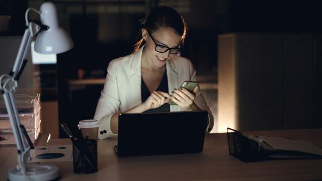 Mulher de óculos e blazer claro, sorrindo, mexe no celular diante de um notebook em um escritório escuro, à noite.