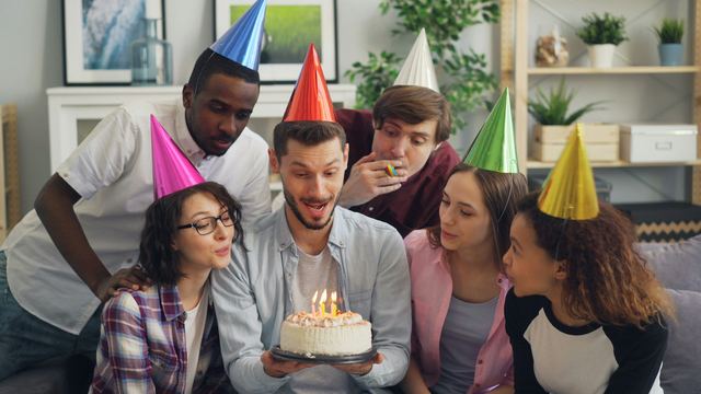 Seis amigos de chapéu de festa em volta de um bolo com velas acesas, um deles prestes a soprar