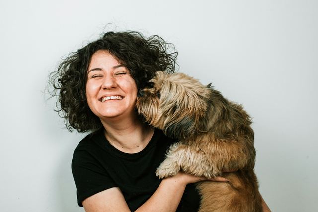 Mulher de cabelo cacheado rindo de olhos fechados enquanto o cachorro peludo, no colo dela, dá um beijo no rosto