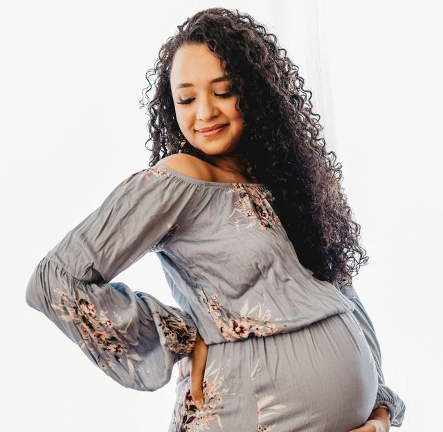 Grávida de cabelo cacheado e vestido cinza florido, sorrindo de olhos baixos com a mão na barriga