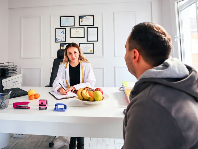 Nutricionista de jaleco sentada em um consultório conversando com um paciente, com uma tigela de frutas sobre a mesa.