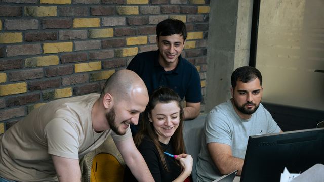 Quatro pessoas em volta de um computador num escritório de parede de tijolos, rindo enquanto trabalham juntas