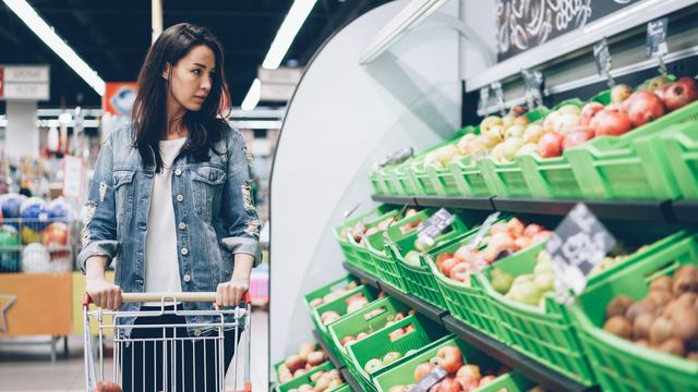 Mulher de cabelo escuro empurrando um carrinho de compras entre as prateleiras de frutas de um supermercado.