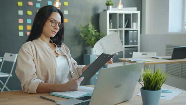 Mulher de blazer conferindo uma pilha de documentos diante do notebook, numa mesa de escritório