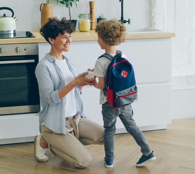 Mãe ajoelhada na cozinha entregando o lanche ao filho de mochila nas costas, os dois sorrindo