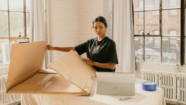 Empreendedora abrindo uma caixa grande diante do notebook, num ateliê de janelões e luz natural