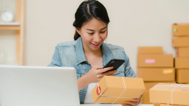 Vendedora sorrindo enquanto confere um pacote com o celular, com notebook e pilhas de caixas atrás