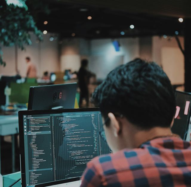 Programador de camisa xadrez, de costas, escrevendo código num monitor num espaço de trabalho compartilhado