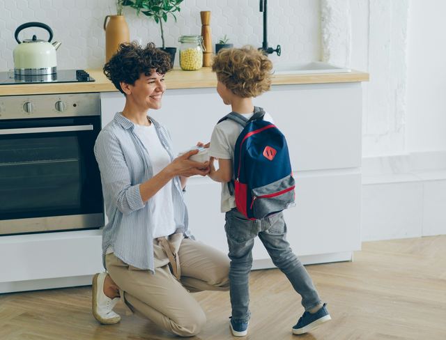 Mãe ajoelhada na cozinha sorri para o filho pequeno, que está de costas com a mochila escolar azul nas costas