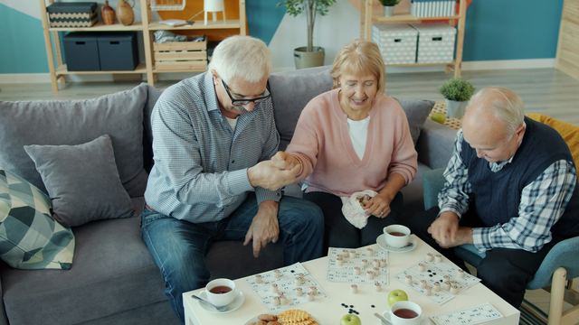 Três idosos sentados num sofá em volta de uma mesa de centro, jogando loto com xícaras de café, sorrindo.