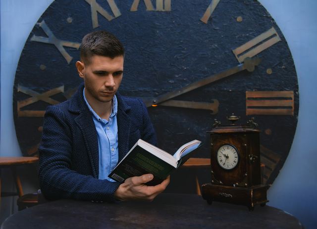 Homem de blazer azul lendo um livro sentado a uma mesa, diante de um grande relógio com numerais romanos ao fundo.