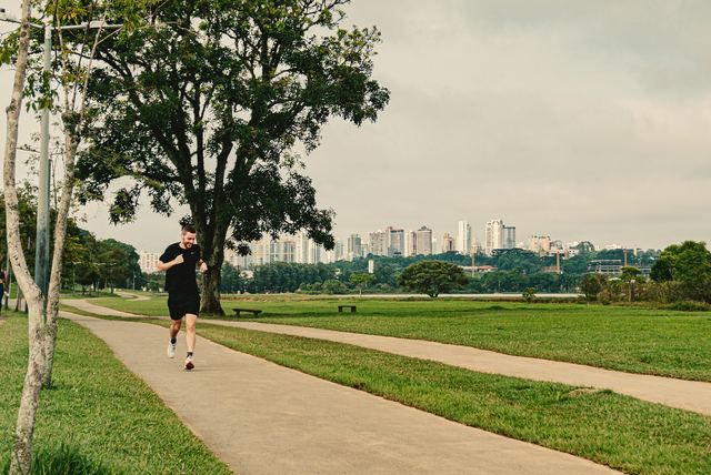 Homem de camiseta e bermuda pretas correndo numa trilha de parque gramado, com prédios da cidade ao fundo