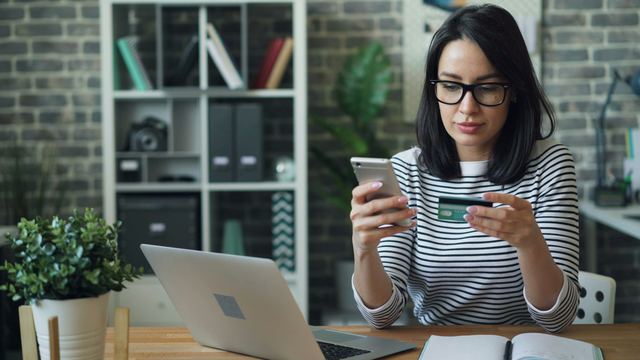 Mulher de óculos e blusa listrada, sentada a uma mesa, segura um cartão de crédito e um celular para fazer um pagamento.