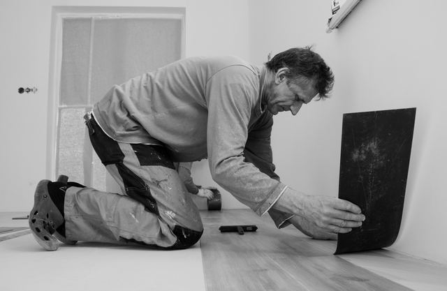 Homem ajoelhado em uma sala assentando uma placa de piso, em foto em preto e branco.