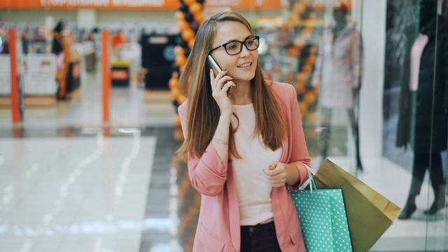 Mulher sorrindo ao telefone, caminhando num shopping com sacolas de papel nas mãos