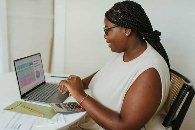 Mulher de óculos e tranças sorri enquanto usa uma calculadora diante de um notebook com gráficos e papéis sobre a mesa.