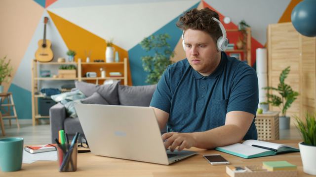 Homem de camisa polo azul e fones de ouvido trabalhando em um notebook, sentado à mesa de uma sala colorida.