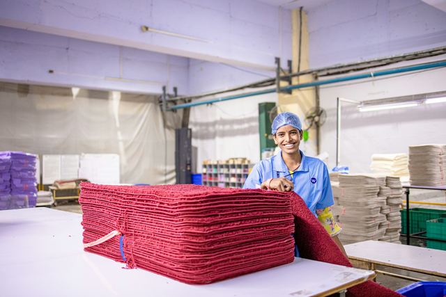 Trabalhadora de touca azul sorri atrás de uma pilha de tecidos vermelhos dobrados em um galpão.