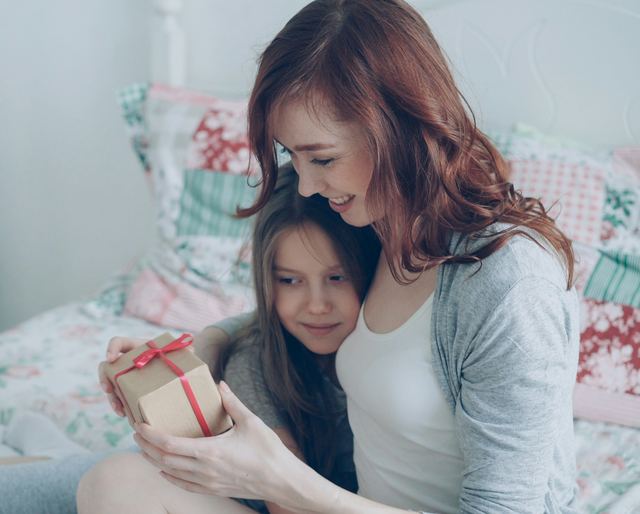 Mãe abraçando a filha sentadas na cama enquanto a menina segura uma caixinha de presente com laço vermelho