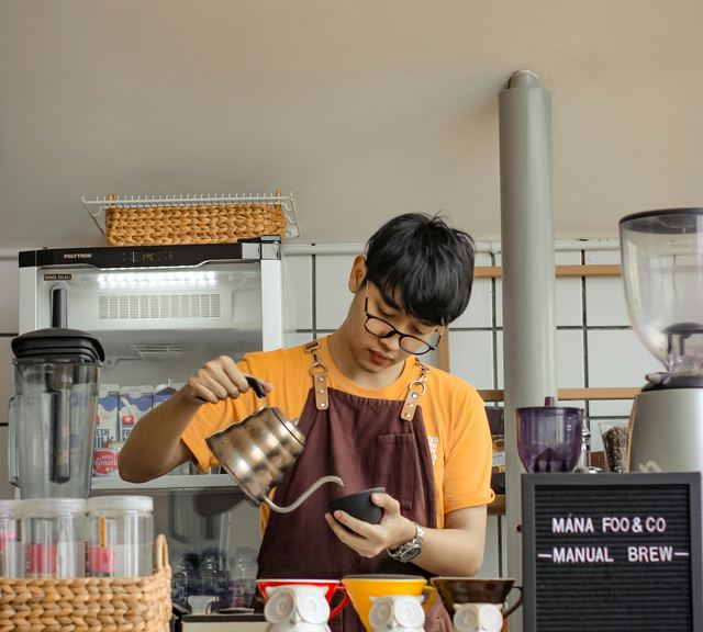 Barista de óculos e avental marrom despejando água da chaleira de bico fino no coador, atrás do balcão