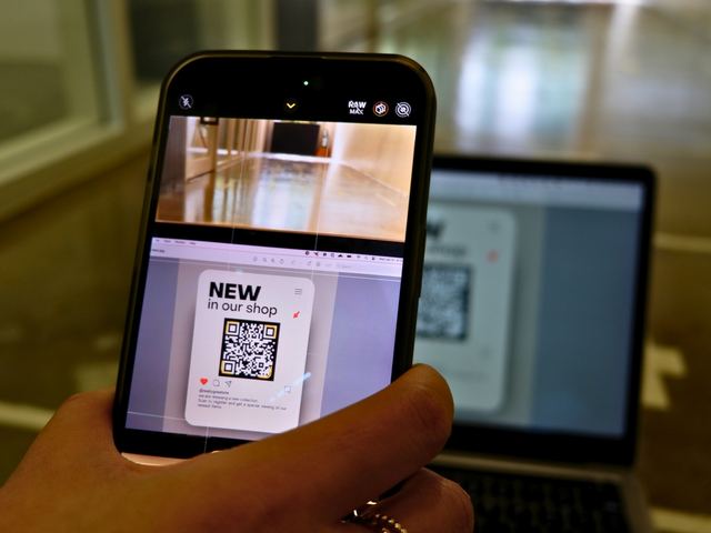 Pessoa apontando o celular para um QR code impresso, diante do notebook, para abrir a lista de produtos de uma loja