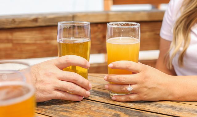 Mãos de duas pessoas segurando copos de cerveja numa mesa de madeira de bar, ao ar livre