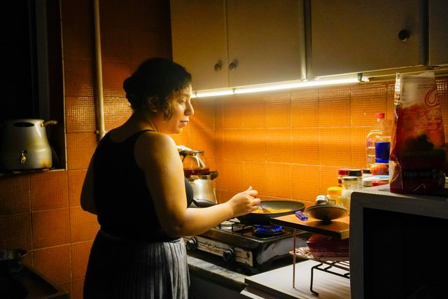 Mulher de perfil cozinhando em uma cozinha pequena de azulejos alaranjados, mexendo uma frigideira sobre o fogão.
